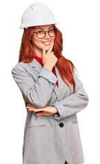 Young redhead woman wearing architect hardhat looking confident at the camera smiling with crossed arms and hand raised on chin. thinking positive.