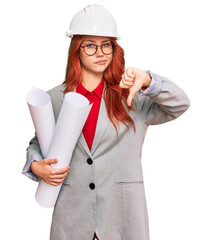 Young redhead architect woman wearing safety helmet holding blueprints with angry face, negative sign showing dislike with thumbs down, rejection concept