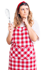 Young caucasian woman wearing apron holding whisk serious face thinking about question with hand on chin, thoughtful about confusing idea