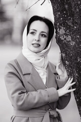 A girl in a light brown coat and a light shawl next to a tree shot on a cloudy April evening in black and white