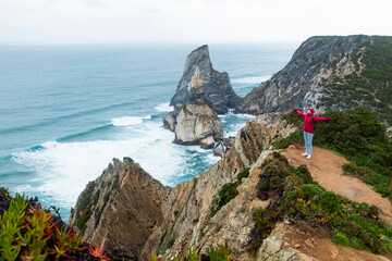 With arms outstretched, a person in a red jacket exudes joy and freedom while standing on a coastal cliff with the vast ocean in the background.