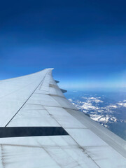 View from airplane window. Wing of an aircraft. Aerial view of blue sky and mountains. The white wing of the plane with clouds below.