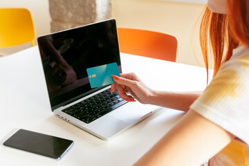 Anonymous redheaded woman is engaged in online shopping, holding a credit card near a laptop while seated at a modern table, highlighting e-commerce convenience