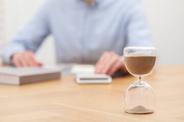 Hourglass with flowing sand on desk. Man using calculator indoors, selective focus