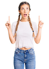 Beautiful caucasian woman with blonde hair wearing braids and white tshirt amazed and surprised looking up and pointing with fingers and raised arms.