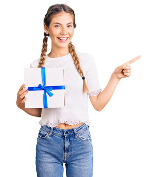 Beautiful caucasian woman with blonde hair holding gift smiling happy pointing with hand and finger to the side