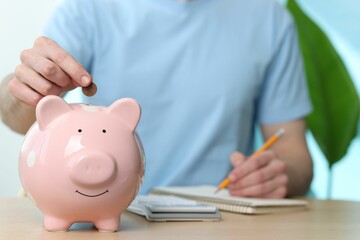 Financial savings. Man putting coin into piggy bank while writing down notes at wooden table, closeup