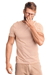 Young caucasian man wearing casual clothes and glasses smelling something stinky and disgusting,...