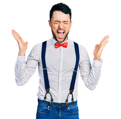Hispanic man with beard wearing hipster look with bow tie and suspenders celebrating mad and crazy...