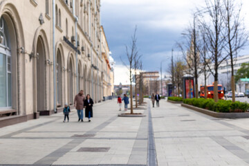 Pedestrian zone of the city street. Defocused photograph of city street. Wide sidewalk with trees and few pedestrians. Spring, cloudy weather