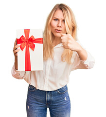 Young beautiful blonde woman holding gift pointing with finger to the camera and to you, confident gesture looking serious