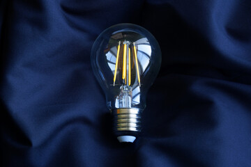Modern quality transparent LED filament lamp with yellow filaments on dark blue silk fabric. Saving electricity. Place for the inscription. Photo. Copy space - obrazy, fototapety, plakaty
