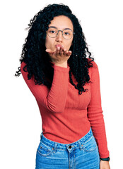 Young hispanic woman with curly hair wearing glasses looking at the camera blowing a kiss with hand on air being lovely and sexy. love expression.