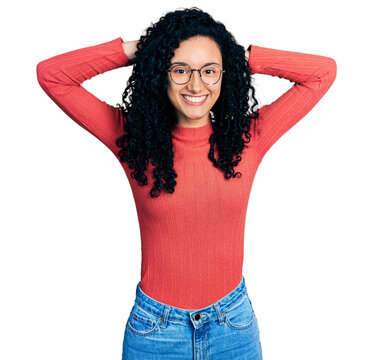 Young hispanic woman with curly hair wearing glasses relaxing and stretching, arms and hands behind head and neck smiling happy