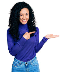 Young hispanic woman with curly hair wearing turtleneck sweater amazed and smiling to the camera while presenting with hand and pointing with finger.