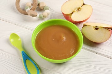 Baby food. Puree of apples in bowl, spoon and toy on white wooden table