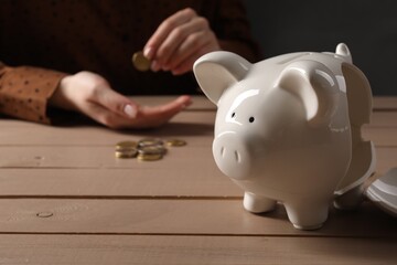Poverty. Woman counting coins at wooden table, focus on broken piggy bank - obrazy, fototapety, plakaty