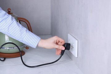 Woman plugging electric kettle into power socket at white table indoors, closeup