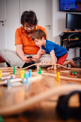 A child and an adult engage with technology and a wooden train set, blending traditional play with modern life in a cozy home environment