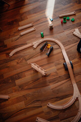 An overhead view of a wooden train track layout, with scattered pieces and toy trains on a warm-toned hardwood floor, awaiting imaginative play
