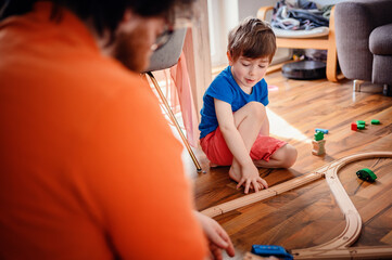 A young boy in blue concentrates on building a wooden toy train track on a hardwood floor,...