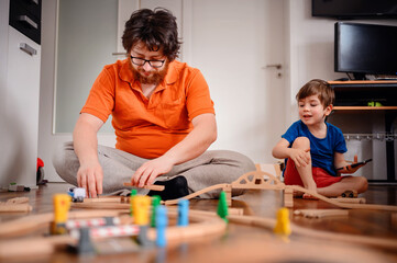 A focused adult builds a wooden train track while a child engages with a smartphone, showcasing the...