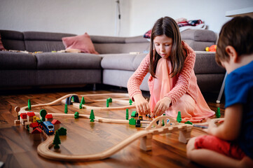 A girl and boy are deeply involved in setting up a sprawling wooden train track, showcasing...