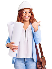 Young latin woman wearing architect hardhat and leather bag holding blueprints pointing thumb up to the side smiling happy with open mouth