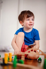 A joyful young boy with a smartphone sits amidst a wooden train setup, his bright laughter echoing...