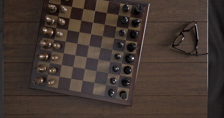 Top view close-up of a chessboard with pieces standing on a wooden table in a room. Glasses lie nearby - 780891693