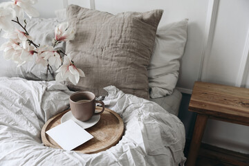 Breakfast in bed concept. Cup of coffee on wooden tray. Blank greeting card, invitation mockup. Bedroom view. Blurred beige linen pillows, blanket. Blooming magnolia branches in vase. Top view.
