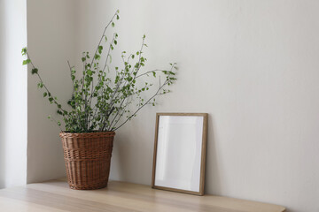 Spring, Easter still life. Elegant Scandinavian living room, home office. Empty vertical wooden picture frame mockup on desk, table. Wicker willow basket with green birch tree branches. Side view.