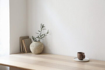 Breakfast, scandi indoor interior still life. Minimal home design concept. Beige ceramic vase with olive tree branches. Cup of coffee, tea on wooden table, desk with old books, empty beige wall mockup
