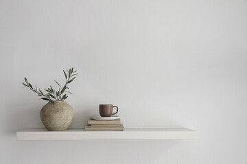 Elegant home still life. Floating shelf. Textured vase with green olive tree branches and old books. Cup of coffee, tea. Modern Mediterranean appartement. White wall background. Interior indoor mockup - obrazy, fototapety, plakaty