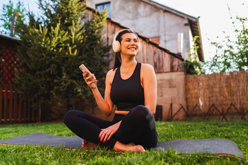 One young girl is stretching and doing yoga on her yoga mat in her backyard while listening to music on wireless headphones and using her mobile phone