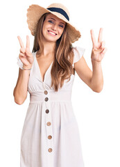 Young beautiful blonde woman wearing summer dress and hat smiling looking to the camera showing fingers doing victory sign. number two.