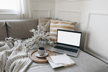 Spring scandinavian living room interior. Sofa with linen yellow striped cushions. Cup of coffee. Cherry plum blossoms in vase. Laptop mockup with blank screen and greeting cards, open book. Home