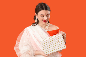Beautiful young Indian woman in sari with gift box on orange background