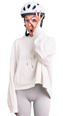 Beautiful brunette young woman wearing bike helmet and sporty clothes doing ok gesture shocked with surprised face, eye looking through fingers. unbelieving expression.