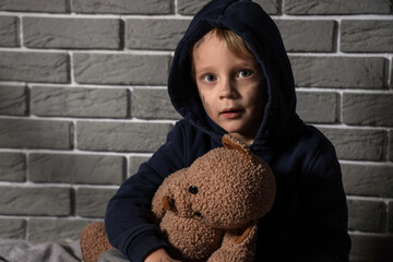 Homeless little boy with toy bear near grey brick wall, closeup