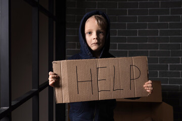 Homeless little boy holding paper with word HELP in dark room