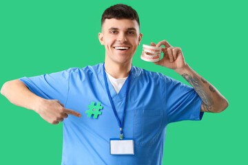 Handsome male dentist with jaw model pointing at hashtag sign on his uniform against green background - Powered by Adobe