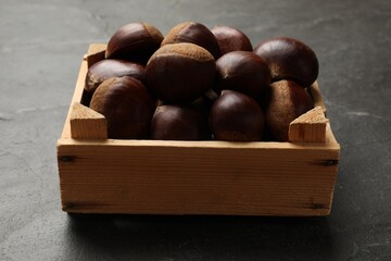 Roasted edible sweet chestnuts in wooden crate on grey textured table, closeup