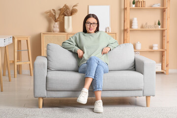 Beautiful young happy Asian woman in stylish eyeglasses sitting on sofa at home