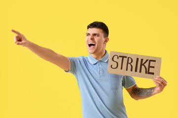 Protesting young man holding placard with word STRIKE pointing at something on yellow background