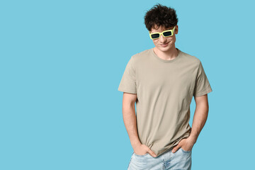 Handsome young man in stylish beige t-shirt and sunglasses on blue background