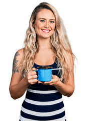 Young blonde woman drinking a cup of coffee looking positive and happy standing and smiling with a confident smile showing teeth