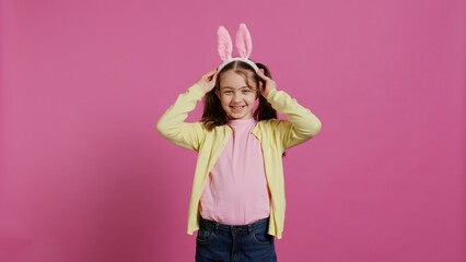 Adorable cute child putting bunny ears and waving at camera, enjoying easter sunday celebration against pink background. Smiling cheery schoolgirl with pigtails saying hello. Camera B.