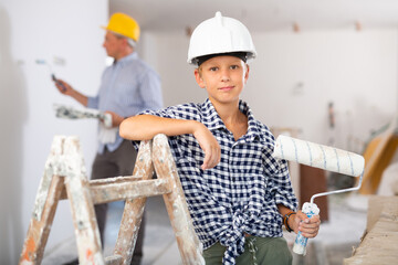 Portrait of positive young boy hardhat holding paint roller. Repair works in apartment.