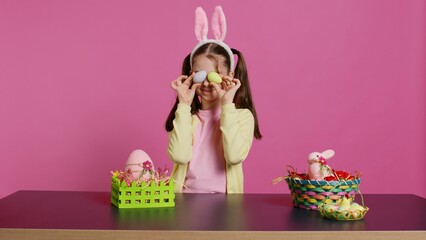 Obraz premium Enthusiastic young girl playing peek a boo in front of camera, using painted easter eggs against pink background. Joyful lovely toddler feeling excited about spring holiday festivity. Camera B.
